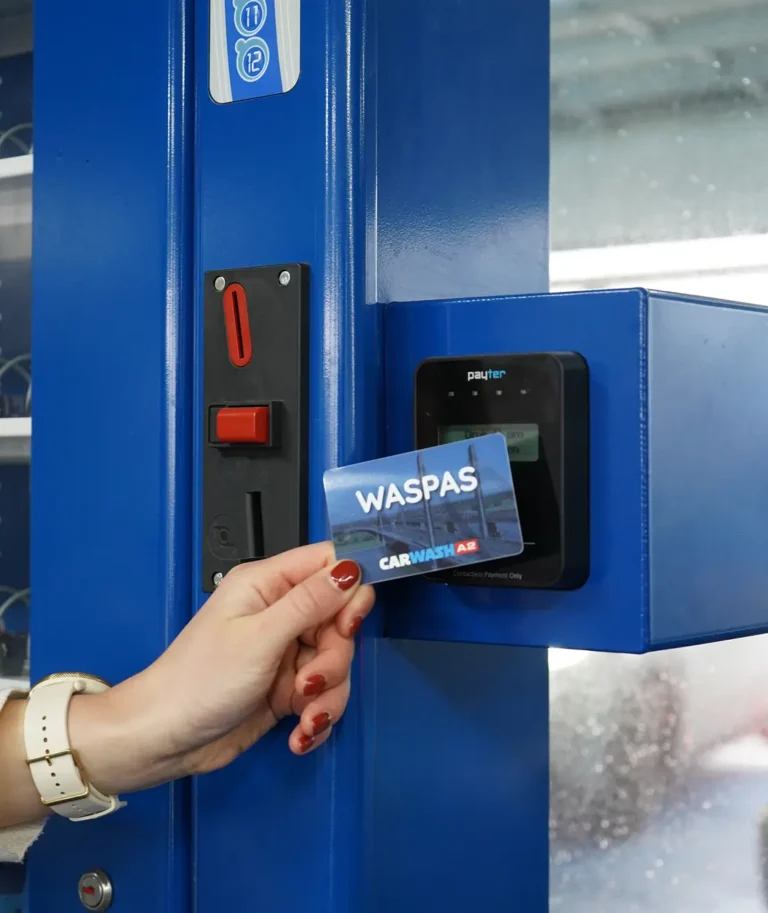 Een hand houdt een Waspas bij een betaalterminal van een autowasstraat.